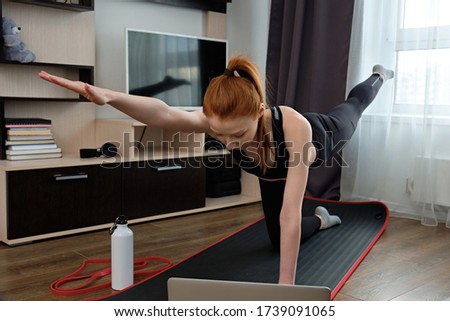 A girl in sportswear performs a sports exercise at home in front of a laptop.