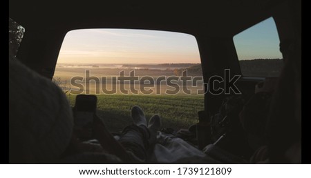 Young woman admiring sunrise in trunk of car van. Girl taking photo on mobile phone against backdrop of nature panorama in fog . View from inside