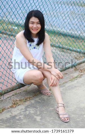 Beautiful asian woman in white dress sitting and smiling and standing near blue iron fence in natural park. 