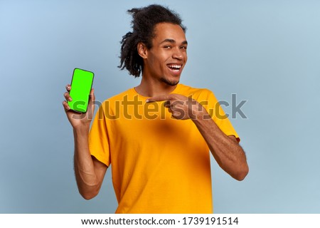 Laughing young african american hipster man points to modern smartphone green screen with alpha channel, isolated on blue background. Green screen advertising space. Gadgets and communication concept