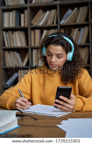 Hispanic teen girl school student wear headphones hold smartphone using distance learning mobile app online watching video course or zoom calling making notes in workbook sit at home or in library. Royalty-Free Stock Photo #1739213303