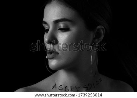 black and white dramatic portrait of a young woman with the inscription aggressive on a black background. the inscription is written in the photographer's hand using mascara ,not a tattoo.