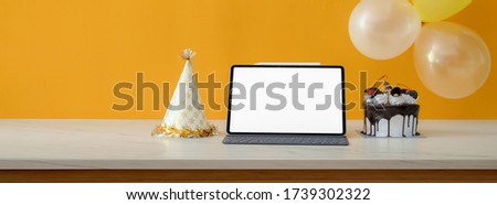 Online birthday party concept with blank screen tablet, cake and party hat on white table with balloon decorated on yellow wall