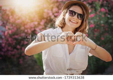 Portrait of a beautiful young stylish woman in a white shirt, hat and sunglasses laughing joyfully, summer mood and vibes, showing heart gesture with hands