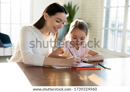 Smiling young mother sit at desk with small preschooler daughter drawing together in album, happy mom or nanny have fun painting picture with little girl child, early development, education concept