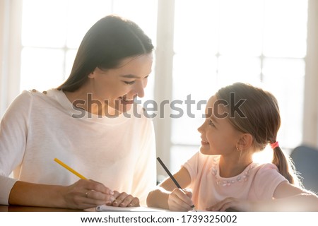 Loving young mother sit at desk studying together with little happy daughter, smiling mom or nanny help teach small girl child, drawing or painting, have fun on weekend at home, education concept