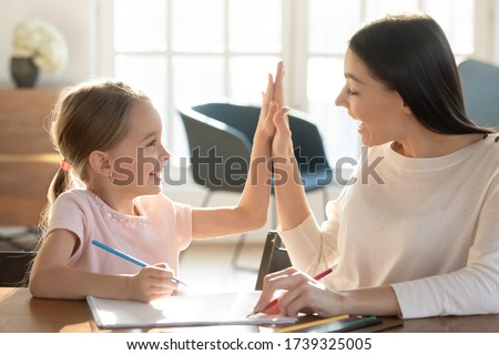 Overjoyed young mom and small daughter give high five celebrate success studying or painting at home together, happy mother or nanny cheering smiling little girl child when drawing, education concept
