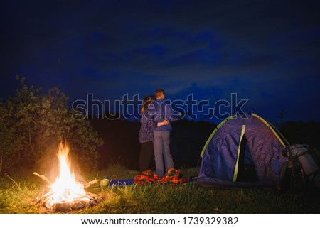 Loving couple hikers enjoying each other, standing by campfire at night under evening sky near trees and tent. Romantic camping near forest in the mountains