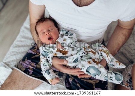 Candid real family moment. Soft focus. Young dark hair father holding his newborn baby son sitting in a bedroom. Boy is wearing body with watercolor animal print. Father's day. Boy is yawning.