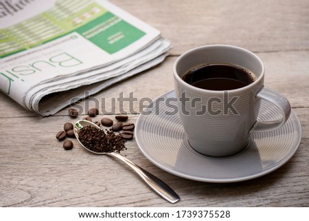 Gray cup of coffee with granulated instant coffee powder in stainless tea spoon with beans and newspaper on wood table background.