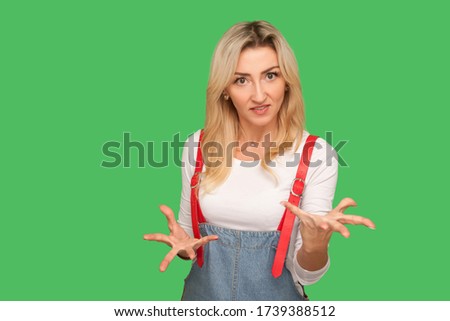 Quarrel, aggressive emotions. Portrait of irritated adult blond woman in stylish denim overalls raising her hands in anger, clenching teeth and looking furious annoyed. indoor studio shot, isolated