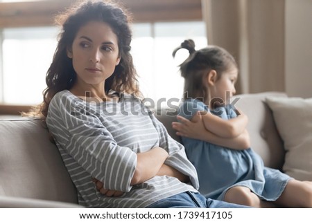 Close up upset annoyed mother and stubborn little daughter sitting on couch with arms crossed, not talking after quarrel, ignoring each other, parent and child, family generations conflict