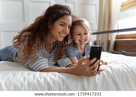 Smiling beautiful mother and adorable little daughter posing for selfie in bedroom close up, lying in bed together, happy young mum holding using phone, watching cartoons with preschool girl