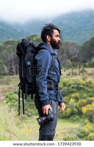 man with long beard, blue jacket and black backpack in nature with a black camera in hand