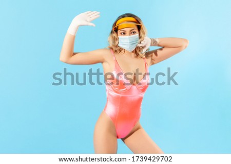 Woman in swimsuit, wearing hygienic face mask and surgical gloves to prevent contagious coronavirus on resort, waving hello with raised hand, welcoming summer vacation with covid-19. studio shot