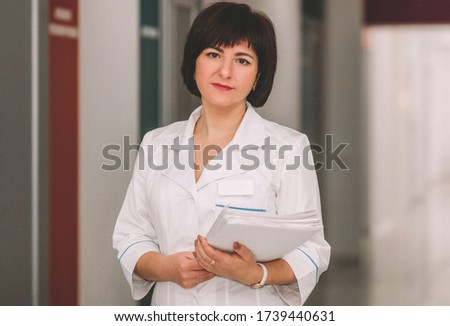 adult female doctor is standing with smile with documents in hands in the corridor of medical clinic . lifestyle medical concept, free space