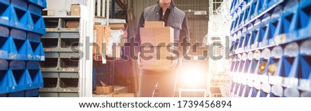 Widescreen image, delivery man in gray uniform carries boxes in his hands at the warehouse. Gold backlight.