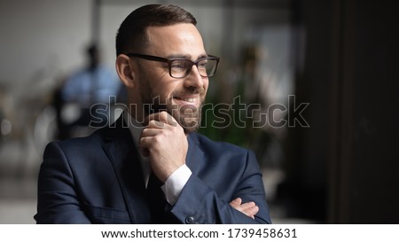 Smiling businessman wearing glasses dreaming about good future close up, touching chin, business vision concept, team leader standing in modern office, looking in distance, thinking about project