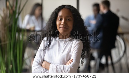 Head shot portrait confident African American businesswoman looking at camera, female team leader posing for photo, standing with arms crossed in modern office with diverse team on background