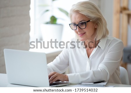 Focused middle-aged businesswoman in glasses sit at desk typing working on laptop, browse wireless internet, concentrated senior female employee consult client online using modern computer gadget Royalty-Free Stock Photo #1739514032