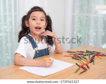 Asian little cute girl sitting and use color pencil drawing cartoon in white paper on wooden table. Preschool lovely kid with  drawing at paper near the window. Learning and education of kid.