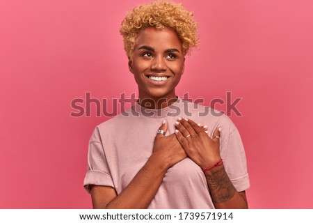 Smiling cheerful dark skinned young female keeps palms on heart, expresses appreciation, pleasure and gratitude, depicts symbol of fight against breast cancer on pink wall. Confession in love concept Royalty-Free Stock Photo #1739571914