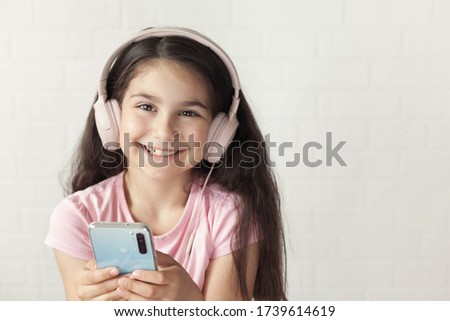 Cute child  girl in headphones is using a smartphone, looking at camera and smiling on light background. Listening music. Online talking. 