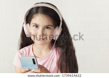 Cute child  girl in headphones is using a smartphone, looking at camera and smiling on light background. Listening music. Online talking. 