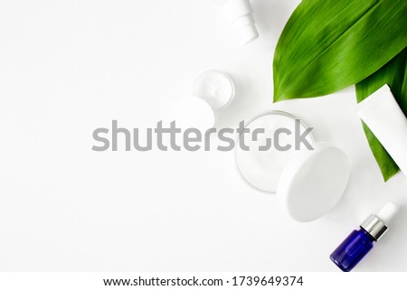 White squeeze tube, bottles of cream, blue dropper glass serum with green tropical leaves flat lay on white background top view copy space. Beauty skincare, natural cosmetic products. Stock photo.