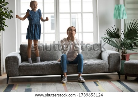 Annoyed tired young Caucasian mother sit on sofa overwhelmed with naughty ill-behaved little daughter jumping playing, exhausted mom bothered by loud noisy small girl child bad behavior at home Royalty-Free Stock Photo #1739653124