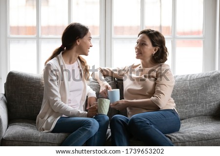 Happy middle-aged mother and adult daughter sit on couch in living room talk drinking tea or coffee, smiling senior mom and grownup millennial girl have fun relax on sofa ta home chatting gossiping Royalty-Free Stock Photo #1739653202