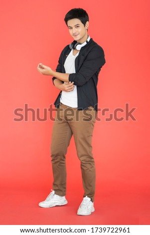 handsome asian confident man doing sleeve button wearing wireless headphone listening to music, joyful happy enjoying sound smiling with happiness lifestyle concept standing red isolated background 