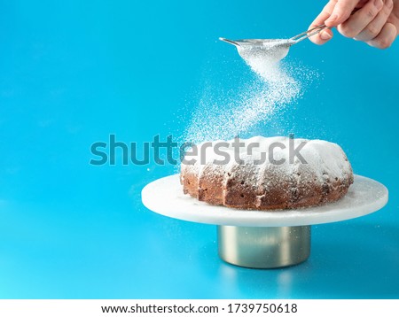 Woman's hand sprinkling icing sugar over fresh home made bundt cake. Powder sugar falls on fresh perfect bunt cake over blue background. Copy space for text. Ideas and recipes for breakfast or dessert Royalty-Free Stock Photo #1739750618