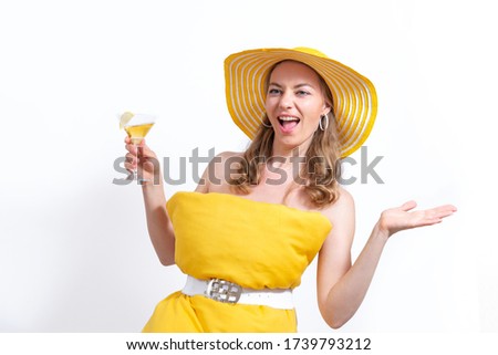 Drunk happy woman in a yellow pillow dress and hat with a glass of cold cocktail smiles on a white background. Summer concept. Pillow Challenge due to stay home isolation. Coronavirus Quarantine