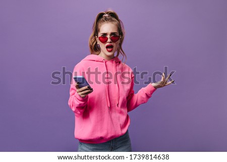 Girl in pink hoodie holding phone and looking into camera with misunderstanding. Pretty woman in street style sweatshirt looks frustrated on purple background