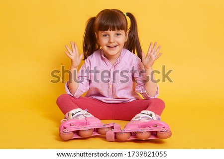 Indoor picture of playful energetic active little girl raising hands, sitting on floor isolated over yellow background, wearing pink casual clothes and roller blades, smiling sincerely. Fun concept.