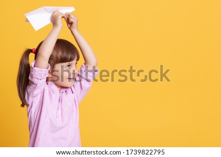 Horizontal picture of playful cheerful kid with ponytails looking aside, having peaceful facial expression, holding paperplane in hands, choosing direction of flight. Copyspace for advertisement.