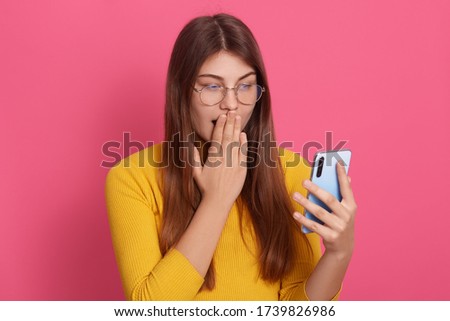 Picture of emotional shocked impressed young model holding smartphone, looking at device screen attentively, covering her mouth with hand, being surprised by news. People and emotions concept.