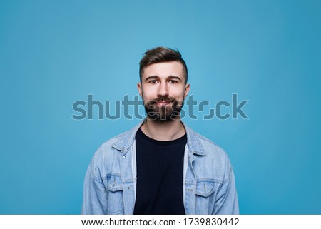 Young cheerful man in black t-shirt and denim jacket. Guy with beard expresses happiness. Blue background.
