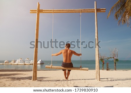 Handsome man swings on sand beach and enjoys landscape under green palms against blue sky backside view
