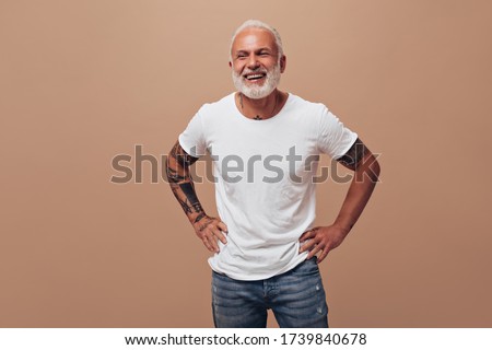 Tattooed man in good mood posing on beige background. Gray-haired guy in white T-shirt and blue jeans laughs into camera Royalty-Free Stock Photo #1739840678