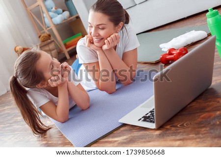 Mother and daughter training online, watching sport fitness program on computer laptop at home. Happy mentoring. Lying on yoga mat, looking at each other, have fun enjoy smile, put hands under chin.
