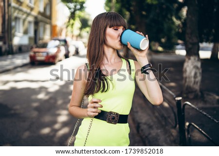 Pretty long hair brunette girl in green light dress with blue paper cup of beverage walking outdoor on the street in the city