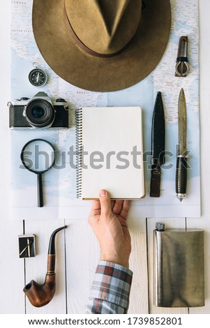 Adventure planning flat lay. Traveler, explorer hand in frame holding empty notepad. Hipster accessories arrangement on map.