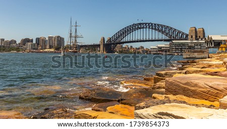 Panoramic view of Sydney harbor bridge and downtown buildings. Sailboat sailing in the harbor towards the bridge. Massive rocks in the foreground
