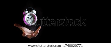 White alarm clock with purple highlights hovering above male hand against black background - narrow banner with copy space