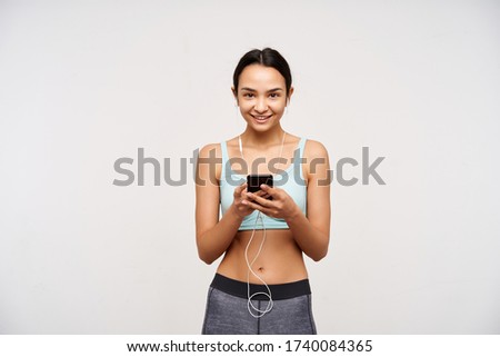 Indoor photo of young pleased brown haired lady without makeup keeping smartphone in raised hands and looking at camera with charming smile, isolated over white background