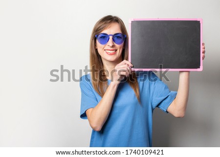 Portrait of a young friendly woman with a smile in a casual blue t-shirt, blue glasses, holding a black chalkboard with blank space for text on an isolated light background. Emotional face