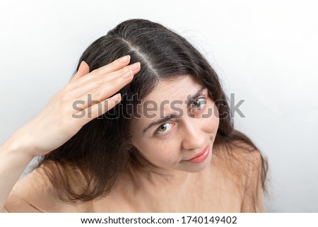 Dandruff and seborrhea. Portrait of a young woman with dark hair, anxiously brushing dandruff from her hair. Gray background. Concept of problems with hair and head lice