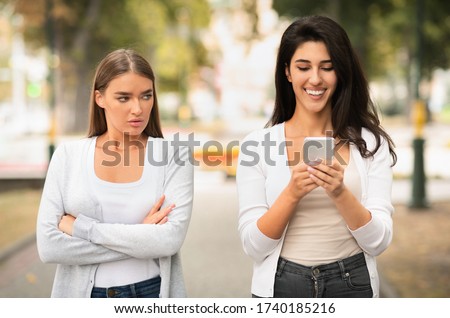 Envy Concept. Jealous Girl Looking At Friend's Phone Walking Together Outdoors At Park. Selective Focus Royalty-Free Stock Photo #1740185216